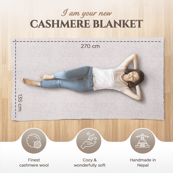 Eine junge Frau liegt entspannt auf einer großen, einfarbig grauen Kaschmirdecke auf hellem Holzfußboden. Die Decke misst 270 cm x 135 cm. Über ihr steht „I am your new Cashmere Blanket“. Am unteren Bildrand sind drei Symbole: ein Wollknäuel mit Text „Finest cashmere wool“, eine Hand mit Feder mit „Cozy & wonderfully soft“ und ein Tempel mit „Handmade in Nepal“. Die Szene wirkt gemütlich und hochwertig.