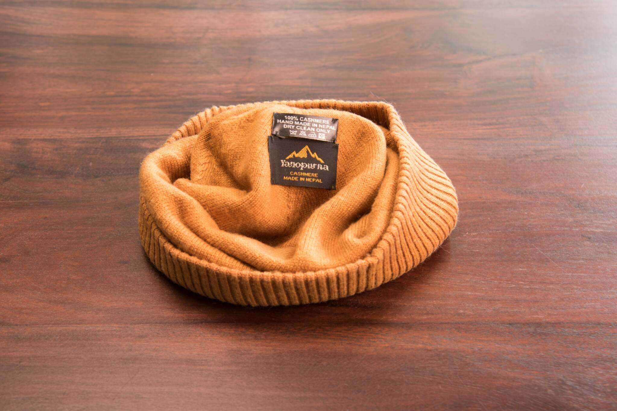 Auf dem Bild ist eine hellbraune Kaschmirmütze mit glatt gestricktem Hauptteil und geripptem Umschlag zu erkennen. Die Mütze liegt auf einer Holzoberfläche. Im Inneren der Mütze sind zwei schwarze Etiketten sichtbar: Auf einem steht „100% Cashmere, Handmade in Nepal, Dry Clean Only“, auf dem anderen befindet sich das Logo „YETOPURNA Cashmere Made in Nepal“ in goldener Schrift. Die Mütze wirkt weich und hochwertig verarbeitet.
